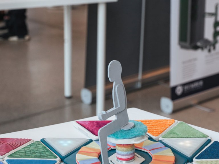 Physical 3D model of person sitting on stool on colorful floor pad design