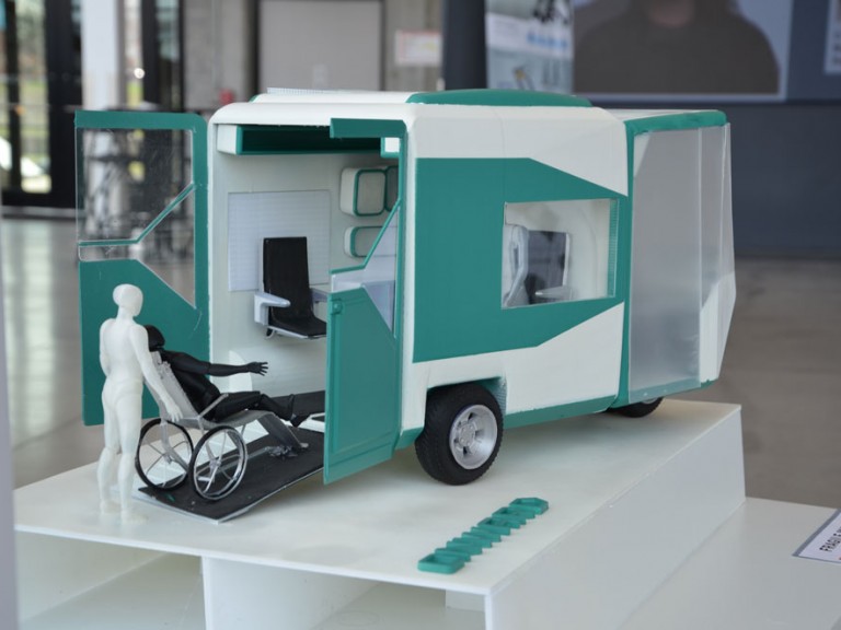 Physical 3D model of one figure rolling another figure in a wheelchair into a transport vehicle
